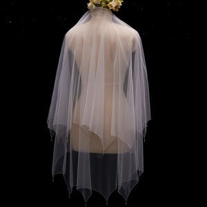 May include: A white, multi-layered tulle wedding veil featuring a handkerchief hemline with delicate beaded edges. The veil is draped over a mannequin and is topped with a small, decorative cluster of cream and pale yellow fabric flowers.