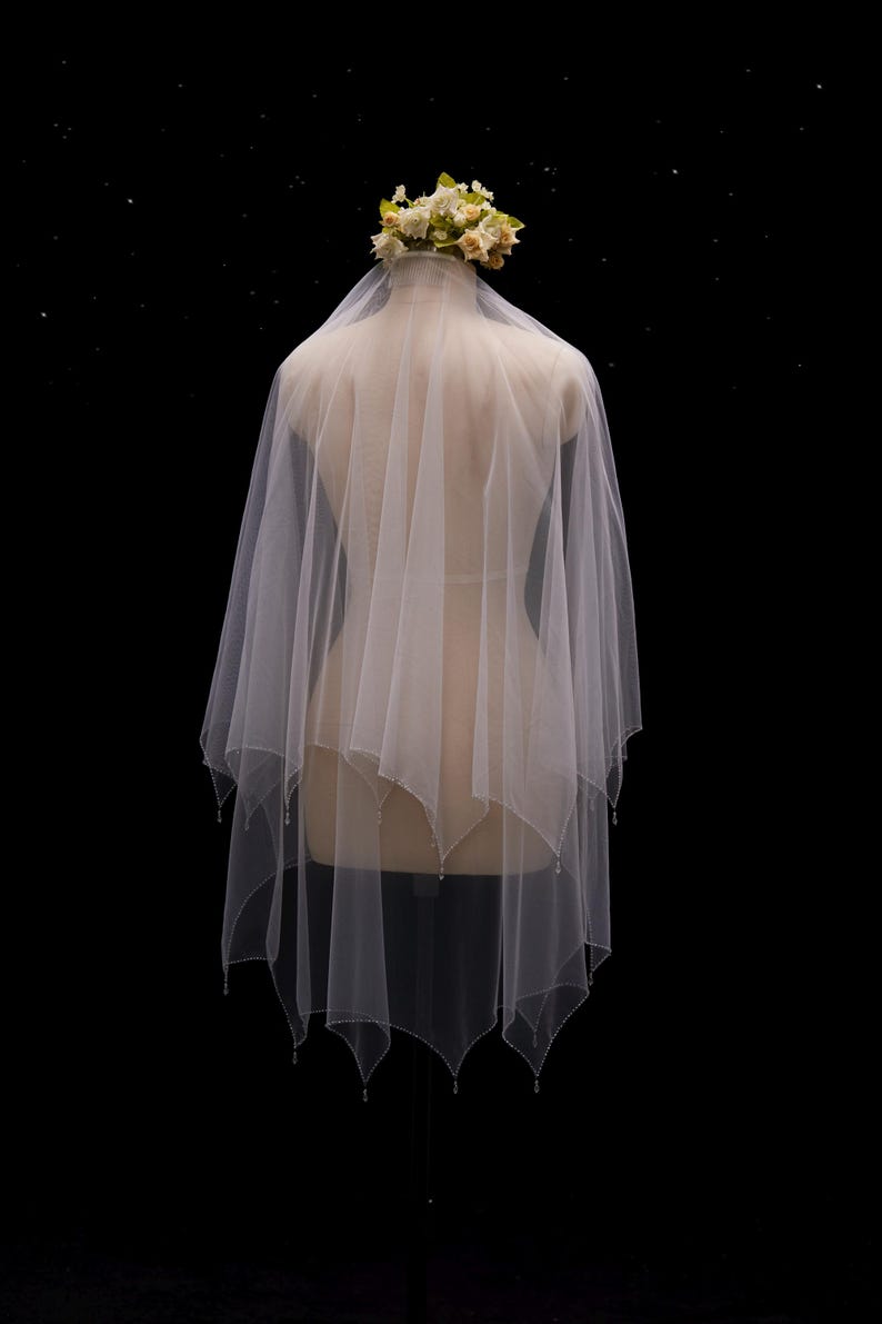 May include: A white, multi-layered tulle wedding veil featuring a handkerchief hemline with delicate beaded edges. The veil is draped over a mannequin and is topped with a small, decorative cluster of cream and pale yellow fabric flowers.
