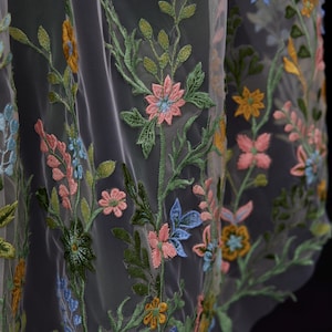 Op de afbeelding: Close-up van transparante stof met geborduurd bloemmotief. De borduursels bevatten roze, blauwe, gele en groene bloemen met groene bladeren. De stof is licht doorschijnend en onthult een donkere achtergrond.