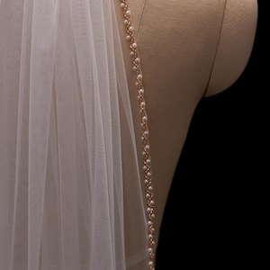 Op de afbeelding: Een doorschijnende, ivoorkleurige bruidssluier met een delicate, met parels bezette rand. De rand heeft kleine, witte parels geregen langs een dunne, roségoudkleurige ketting. De sluier is over een mannequin gedrapeerd, met een zwarte achtergrond.