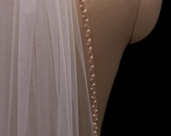 May include: An ivory bridal veil with a beaded trim. The trim is made of small, white pearls strung along a rose gold-coloured chain. The veil is draped over a mannequin, against a black background. The veil is sheer.