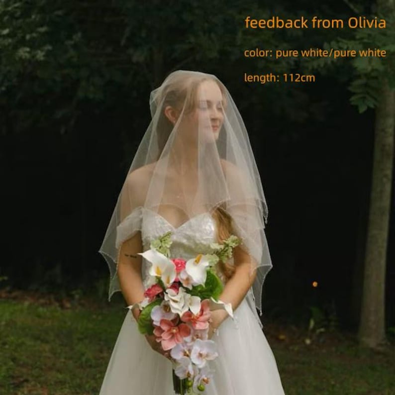 May include: A pure white wedding veil measuring 112 cm in length, draped over the head. The veil is made of sheer, lightweight fabric and is paired with a white dress and a bouquet of white and pink flowers. Text reads: feedback from Olivia.