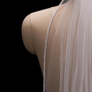 May include: A bridal veil with a delicate beaded edge and sheer, flowing fabric. The veil is attached to a mannequin, showcasing its design and drape. The background is black, highlighting the veil's details.