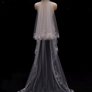 May include: A white bridal veil with floral lace detailing and a floral headpiece. The veil is displayed on a mannequin against a black background. The veil has a two-tiered design with a long, flowing train.
