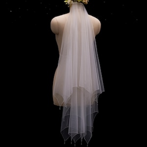 May include: A two-tier white tulle wedding veil featuring a scalloped edge with delicate bead embellishments along the hem. The veil is attached to a floral headpiece composed of cream and pale green fabric flowers, secured on a mannequin.