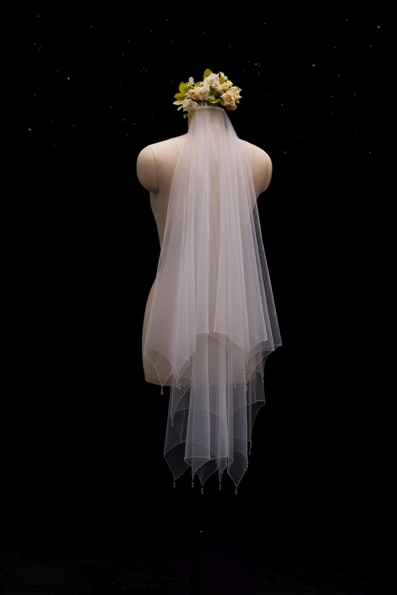 May include: A two-tier white tulle wedding veil featuring a scalloped edge with delicate bead embellishments along the hem. The veil is attached to a floral headpiece composed of cream and pale green fabric flowers, secured on a mannequin.