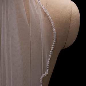 May include: A sheer white bridal veil with a delicate beaded edge. The beads are clear and small, adding subtle sparkle. The veil is draped over a mannequin, showcasing its flowing fabric and design.