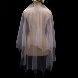 May include: A white, multi-layered tulle veil featuring a scalloped hemline adorned with small, clear beads at each point. The veil is draped over a mannequin and topped with a cluster of cream and pale green artificial flowers.