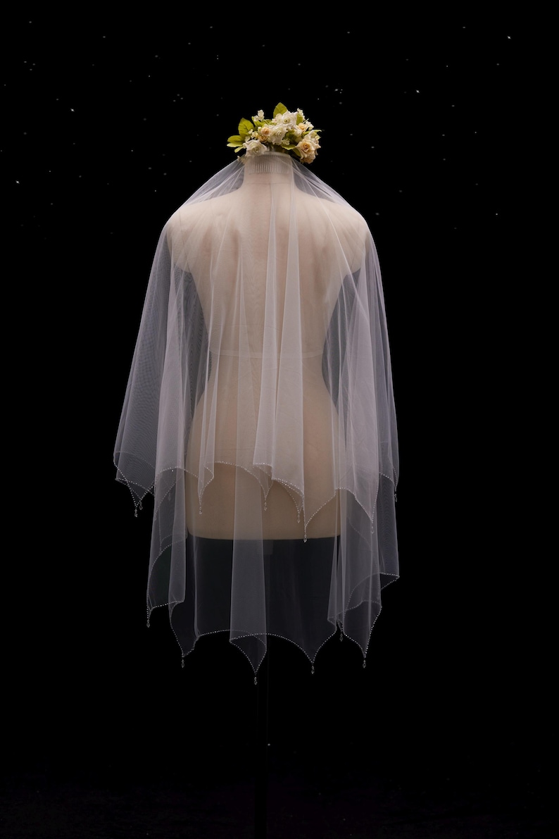 May include: A white, multi-layered tulle veil featuring a scalloped hemline adorned with small, clear beads at each point. The veil is draped over a mannequin and topped with a cluster of cream and pale green artificial flowers.