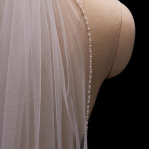May include: A white, sheer bridal veil with a beaded edge. The veil is draped over a mannequin, highlighting its soft texture and elegant design. The beads are small and clear, adding a subtle sparkle to the fabric.