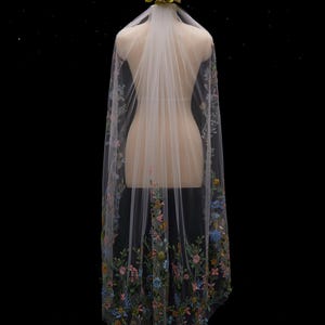 May include: A sheer white wedding veil with floral embroidery in shades of pink, blue, yellow, and green. The veil is displayed on a mannequin and is adorned with a floral headpiece. The veil cascades down the back.