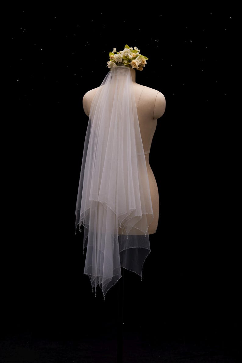 May include: A two-tier white tulle wedding veil featuring a scalloped edge with delicate bead accents. The veil is attached to a floral headpiece composed of small cream and pale yellow fabric flowers, displayed on a mannequin torso.