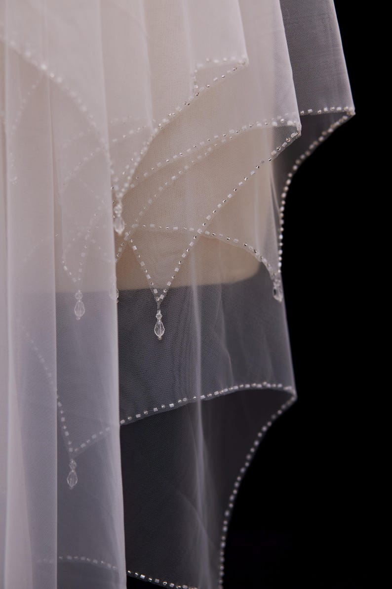 May include: A sheer, off-white bridal veil featuring multiple layers of soft tulle fabric. The edges are adorned with intricate silver beadwork arranged in a scalloped pattern, accented by small, clear, teardrop-shaped crystal pendants.