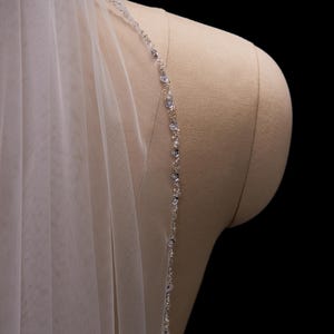 Puede incluir: Un velo de novia marfil transparente con un delicado ribete de cuentas. El ribete presenta pequeños cristales transparentes y detalles florales. El velo está drapeado sobre un maniquí.