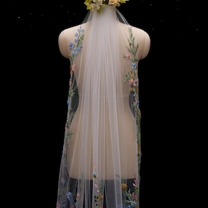 Op de afbeelding: Een doorschijnende witte bruidssluier met kleurrijke bloemenborduurwerk langs de randen. De sluier wordt op een mannequin getoond en is versierd met een bloemenkrans. Het borduurwerk bevat blauwe, roze en gele bloemen.