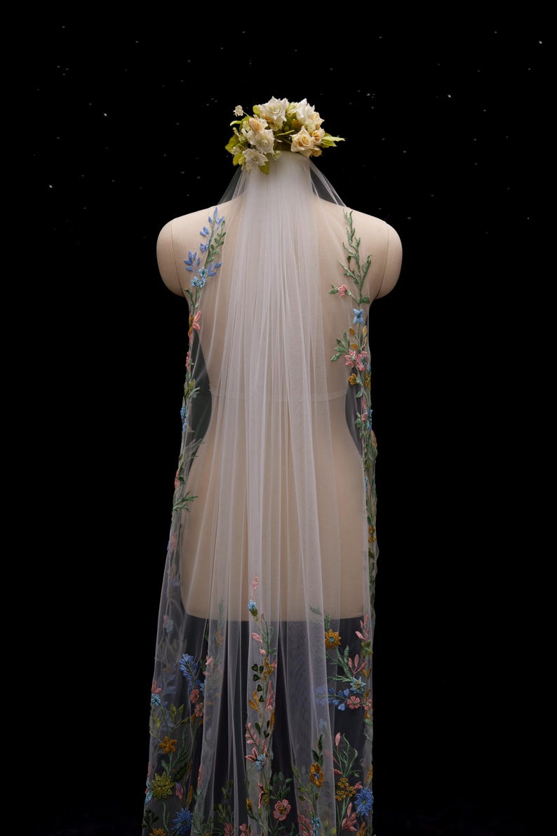 Op de afbeelding: Een doorschijnende witte bruidssluier met kleurrijke bloemenborduurwerk langs de randen. De sluier wordt op een mannequin getoond en is versierd met een bloemenkrans. Het borduurwerk bevat blauwe, roze en gele bloemen.