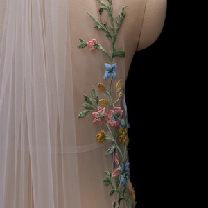 Op de afbeelding: Een doorschijnende, ivoorkleurige sluier met delicate bloemenborduurwerk. Het borduurwerk toont een verscheidenheid aan kleurrijke bloemen en groene bladeren, wat een elegant ontwerp cre&euml;ert. De sluier is over een mannequin gedrapeerd.