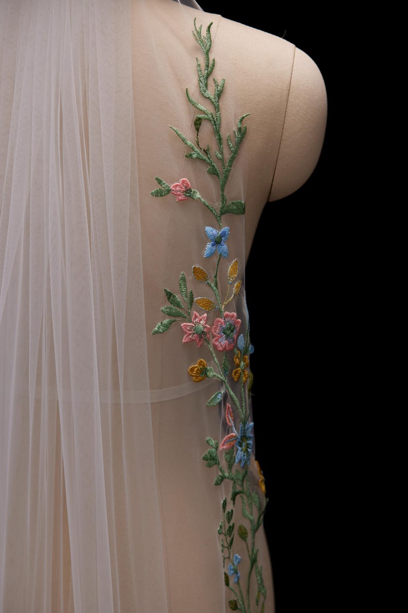 Op de afbeelding: Een doorschijnende, ivoorkleurige sluier met delicate bloemenborduurwerk. Het borduurwerk toont een verscheidenheid aan kleurrijke bloemen en groene bladeren, wat een elegant ontwerp cre&euml;ert. De sluier is over een mannequin gedrapeerd.