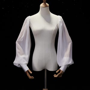 May include: A white blouse featuring long, voluminous bishop sleeves made of a sheer, lightweight fabric. The garment has fitted cuffs at the wrists and is displayed on a mannequin torso against a dark background to highlight the sleeve structure.