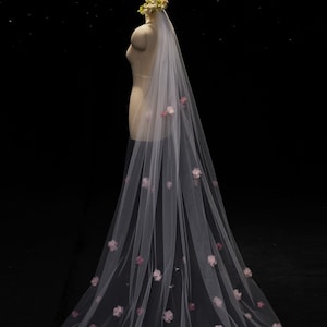 May include: A long, floor-length white tulle wedding veil draped over a mannequin. The veil features scattered, three-dimensional pink floral appliqués throughout the fabric and a cluster of white and green floral accents at the crown.