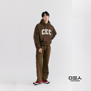May include: A person wearing a brown hoodie with the letters "CEC" in white, paired with brown pants and red and black sneakers. The outfit is set against a white backdrop, showcasing a casual streetwear style.