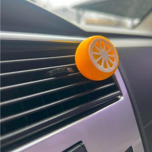 May include: A bright orange car air freshener shaped like a citrus fruit slice, with a white interior design. The air freshener is attached to a car vent, with a black and silver dashboard in the background.
