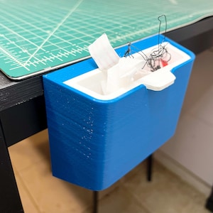 May include: A blue, rectangular tissue box holder with a white interior, mounted on a black table. The holder is made of a solid material and has a curved design. White tissues are visible inside the holder.