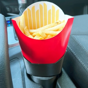 May include: A red and yellow paper container filled with golden french fries, placed in a black car cup holder. The container has a red exterior and a yellow and white interior design.
