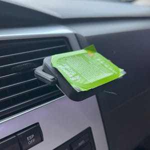 May include: A black 3D-printed car air vent clip holding a green and white packet of Sweet 'N Sour sauce. The packet is rectangular with text and sits in front of a car's air vent.
