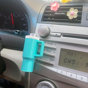 May include: A turquoise blue car air freshener with a white top and a handle. The air freshener is hanging from the air vent in a car. The car radio is visible in the background with the text "FM 1 CH5" displayed.