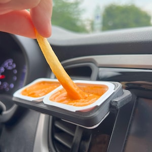 May include: A black 3D-printed car vent sauce holder with two white sauce containers filled with orange sauce. A french fry is dipped into the sauce. The holder is attached to a car's air vent.