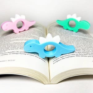 May include: Four colorful dinosaur-shaped book page holders in pink, green, and blue, with white accents. The book page holders are resting on the pages of an open book.