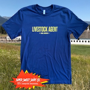 Op de afbeelding: Een koninklijk blauw T-shirt met de tekst "LIVESTOCK AGENT * DARBY, MONTANA *" in geel. Het shirt is gefotografeerd tegen een groene grasveld met een witte schuur op de achtergrond.