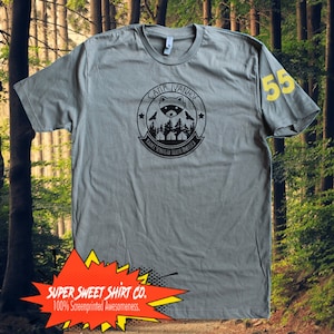 May include: A gray t-shirt with a black and white graphic of a raccoon in a circle with the text "Camp Ivanhoe" and "Boy Scouts of North America". The number 55 is printed in yellow on the right sleeve.
