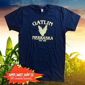 Könnte beinhalten: Ein marineblaues T-Shirt mit einer gelben Grafik von zwei Maiskolben und dem Text "GATLIN NEBRASKA POP. 968". Das Shirt hat ein weißes Etikett mit dem Text "SUPER SWEET SHIRT CO. 100% Screenprinted Awesomeness."