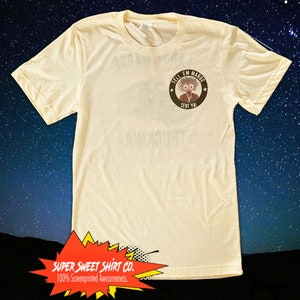 May include: A light yellow t-shirt featuring a black and white cartoon character graphic on the front. The graphic is a circle with the text "TELL 'EM MARGE SENT YA" and a cartoon character with wild hair and a surprised expression. The shirt is from "SUPER SWEET SHIRT CO. 100% Screenprinted Awesomeness."