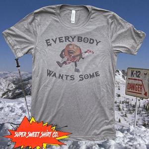 Op de afbeelding: Een grijs T-shirt met een cartoonfiguur dat gitaar speelt. De tekst "EVERYBODY WANTS SOME" staat op het shirt gedrukt. Het shirt is op een besneeuwde achtergrond met een "K-12 DANGER" bord.