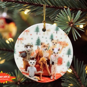 May include: Round ceramic Christmas ornament featuring a whimsical illustration of anthropomorphic animals in a cozy setting. The ornament has a gold string for hanging and is decorated with a festive pattern of trees and snowflakes.