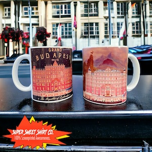 May include: Two white ceramic mugs with wraparound designs. One mug features the text "GRAND BUDAPEST HOTEL" over a detailed illustration of a building. The other mug displays a pink building with a mountain backdrop. The mugs are screenprinted.