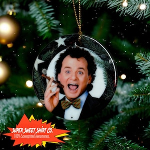 Może przedstawiać: Okrągła ozdoba choinkowa przedstawiająca portret mężczyzny o ciemnych włosach, ubranego w smoking i muszkę. Trzyma w prawej ręce cygaro i ma zaskoczony wyraz twarzy. Ozdoba jest ozdobiona zieloną gałązką sosny i białą gwiazdą.