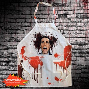 Puede incluir: Delantal blanco con un gráfico de la cara de un hombre salpicada de pintura roja y salpicaduras de pintura roja adicionales. El delantal tiene un lazo para el cuello y lazos en la cintura. La esquina inferior izquierda tiene un logotipo que dice "SUPER SWEET SHIRT CO. 100% Screenprinted Awesomeness."