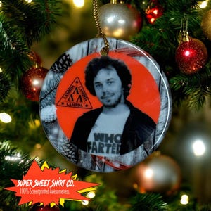 May include: A round ceramic ornament with a woodgrain background. The ornament features a photo of a person with a black shirt and a red circle with a triangle and the word "Lambda" written three times. The text "Who Farted" is written on the shirt.