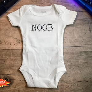 May include: White baby onesie with the word "NOOB" printed in black capital letters. The onesie has short sleeves and a round neckline. The onesie is on a wooden surface, with a keyboard and game controller in the background.