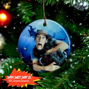Puede incluir: Un adorno navideño redondo de cerámica con un hombre con una expresión de sorpresa sosteniendo un pequeño árbol de Navidad. El adorno es azul con copos de nieve blancos y una cuerda dorada.