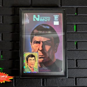 May include: Framed comic book cover featuring Leonard Nimoy, with a smaller trading card of Nimoy below. The comic cover has a purple background with Nimoy's portrait. The frame is black, and the background is a black brick wall.