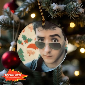 Op de afbeelding: Ronde kerstversiering met een portret van een man met een zonnebril, versierd met een feestelijk kerstman- en hulstpatroon. De versiering hangt aan een gouden draad, met een kerstboom op de achtergrond. De tekst "SUPER SWEET SHIRT CO." is zichtbaar.