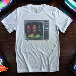 May include: White t-shirt with a retro television graphic showing three figures wearing sunglasses. The graphic is in a vintage style. The shirt is made of a soft material. The shirt has a crew neck and short sleeves.