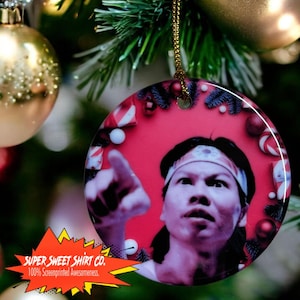 May include: A round ceramic Christmas ornament featuring a man with a headband pointing upwards. The ornament has a red background with white and red candy canes and other Christmas decorations. The text "SUPER SWEET SHIRT CO. 100% Screenprinted Awesomeness." is printed on the bottom of the ornament.