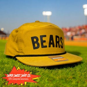 May include: A mustard yellow baseball cap with a black adjustable strap and a black "BEARS" logo on the front. The cap is sitting on a green grassy surface.