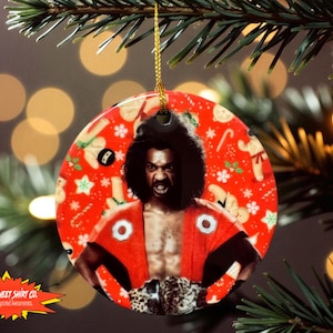 May include: Round Christmas ornament featuring a man in a red robe with white circles and a leopard print belt. The background is red with Christmas-themed illustrations. The ornament is hanging from a gold string.